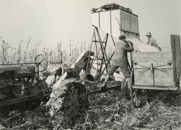 McCormick-Deering power drive corn picker in action in the field. Two men are emptying the contents from the picker into a wooden wagon.