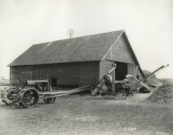 Farmall Regular tractor powering a McCormick Deering No. 2 corn sheller near a barn.