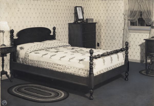 A bed covered with a decorative bedspread is standing in the middle of a bedroom, surrounded by an end table with a lamp and a dresser. What appears to be a dressing table is standing to the right near a curtained window. Braided rugs are lying on the floor beside the bed and beneath the dressing table.