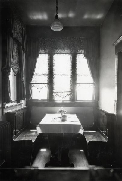 Two benches, and a table covered with a cloth is set with a vase of flowers and a creamer dish, are standing beneath curtained windows in a farmhouse breakfast nook.