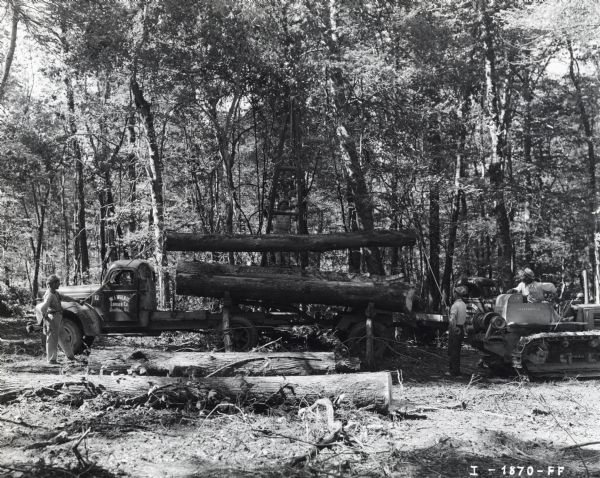 A group of men load cut logs onto the back of an International truck in a wooded area.  The writing on the truck cab reads: "W.I. Wilkie Lumber Co. Smithton, Ark." An International crawler tractor (TracTracTor) is on the right.