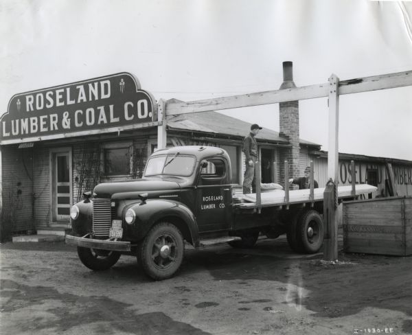 A man stands on the bed of an International K-5 truck loaded with cut lumber while another man stands nearby. The Roseland Lumber & Coal Company building is behind them.
