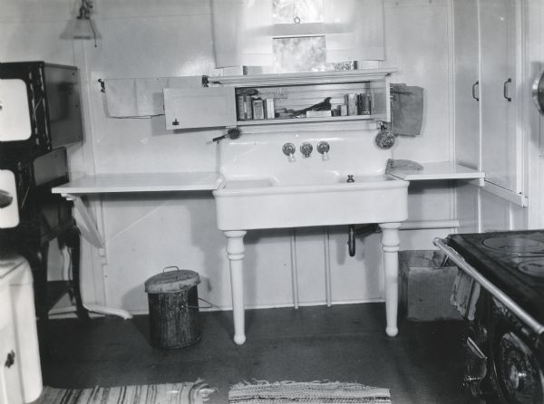 A sink standing against a wall inside a farmhouse kitchen. A stove is to the right of the sink, and a garbage bin stands beneath it.