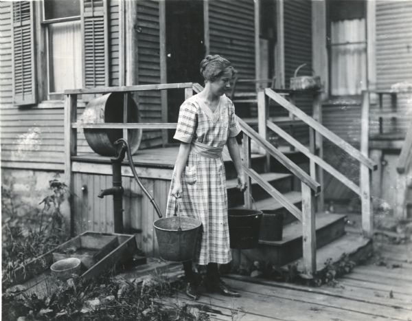 A woman carrying two metal buckets full of water away from a well pump near the porch of a farm house.
