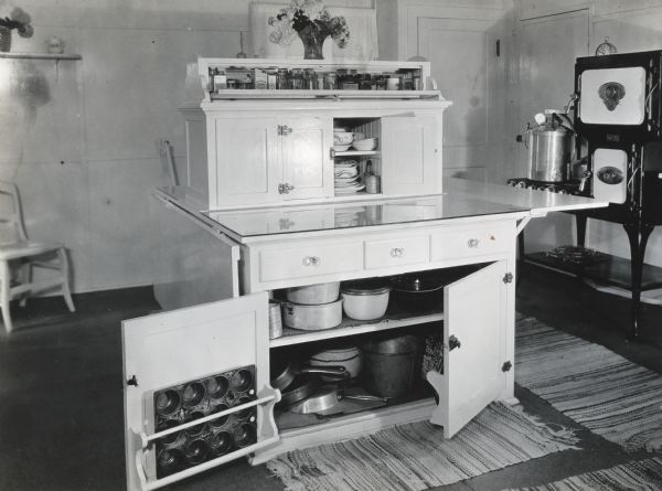 A table with built-in cabinets and shelf space standing in the middle of a farmhouse kitchen, next to a wooden chair to the left and a stove and ovens to the right.