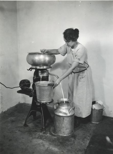 Mrs. Waggoner steadying a metal pail on the platform of a cream separator as it is collecting cream at International Harvester's demonstration farm. Milk is flowing into a metal milk can on the floor.