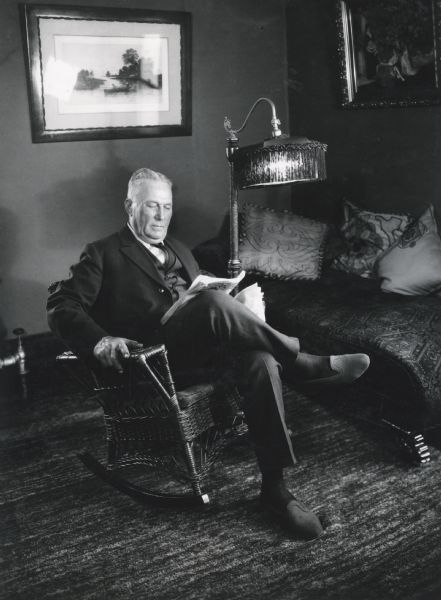 Mr. Lutfring sitting in a wicker rocking chair while reading a newspaper. The room is furnished with an upholstered divan, lamp with fringe shade, carpeting, and two framed paintings are hanging on the walls.