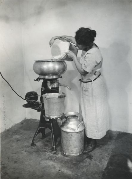 Mrs. Waggoner straining milk from a bucket into an electric cream separator using a piece of cheesecloth. A metal bucket is standing on a platform and a metal milk can is resting on the floor.