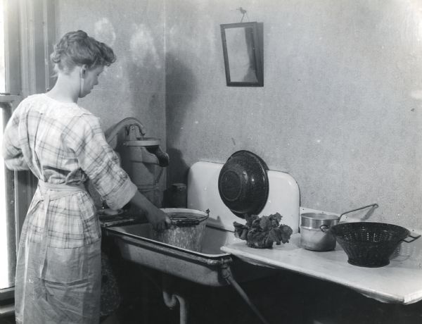 A woman filling a bucket with water from an indoor well pump while standing at a kitchen sink. A mirror is hanging on the wall and a potted plant, canister, and colander are sitting on a counter next to the sink.