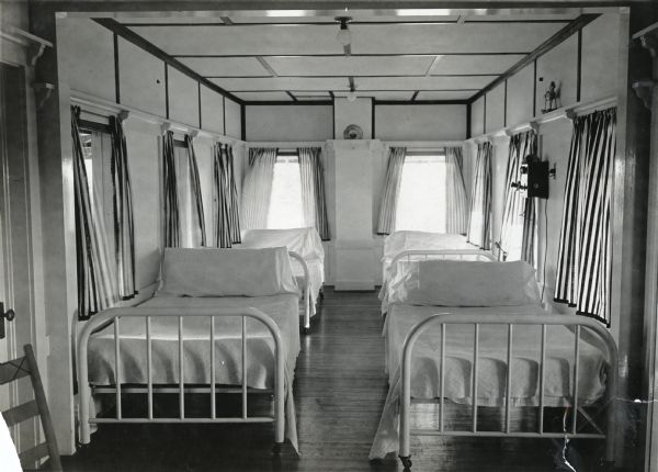 Four beds line either side of the sleeping porch at the farmhouse of J.E. Joyce. A telephone is hanging on the wall and striped curtains are covering the opened windows.