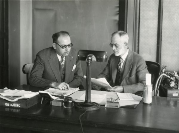 Professor Perry G. Holden (right) and George W. Anstrand sitting behind a desk and looking at papers.