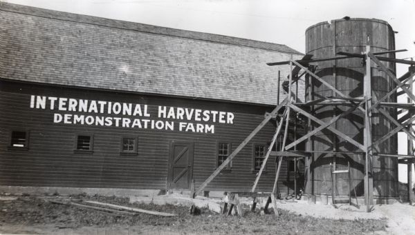 Wooden scaffolding surrounds a silo on an International Harvester demonstration farm. A barn stands next to the silo, painted with a sign that says: "International Harvester Demonstration Farm".