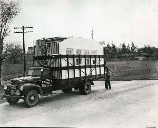 A man loading an International K-5 truck with sectional housing pieces and lumber while parked alongside a rural road. The text on the truck reads: "Economy Portable Houses. West Chicago, Ill. J.L. Lynch. Field Rep. Box 177, Phone 2. Boswell, Ind." Several farmhouses and a windmill are in the background.
