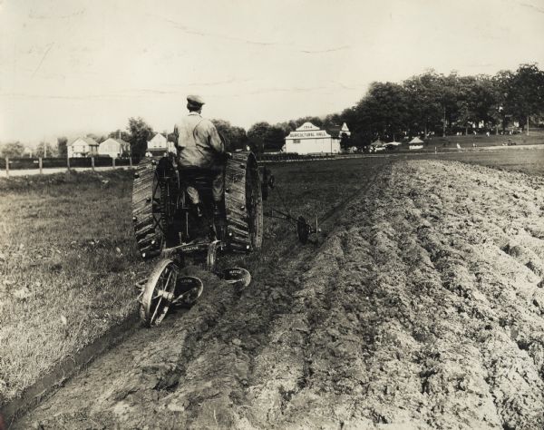Rear view of an Oliver No.84 two row plow in field. In the background is a building with a sign that says: "1914 Agricultural Hall."
