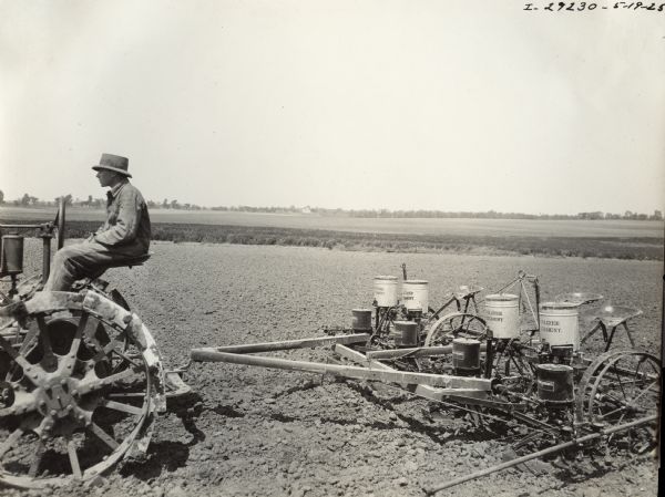 Side view of man driving a Farmall regular pulling a McCormick-Deering corn planter and fertilizer attachment in a field.