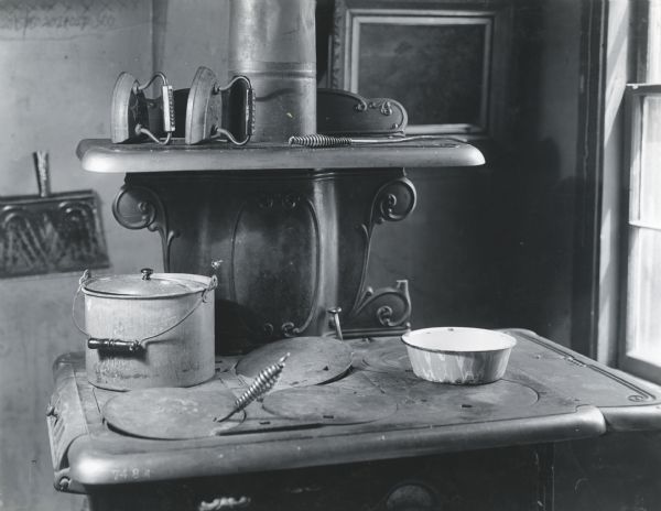 Pots, kitchen utensils, and two irons sitting on the stove of a wood-burning oven. A framed painting and a dustpan hang on the wall in the background.