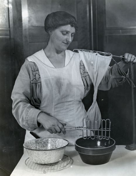 A woman wearing an apron and a hairnet standing at a counter while using a hoop strainer to make cheese(?). She is using another metal utensil to press whey from the curd, held in a piece of cheesecloth, into a bowl.