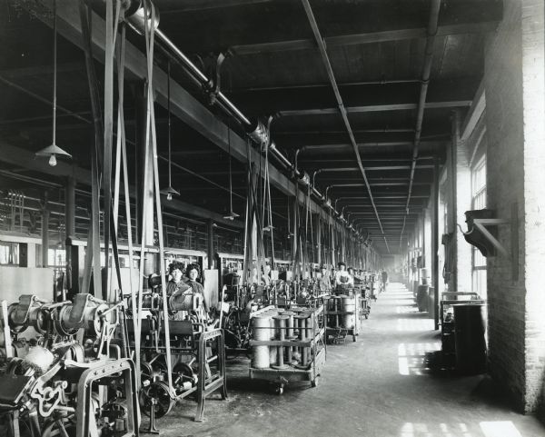 Male and female factory workers wear protective hair bonnets while working in a room where twine is wound, weighed, and inspected. The workers stand in a row of belt-driven machines near a row of windows. The location may be a twine mill at International Harvester's Deering Works.