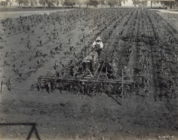 Elevated view of man operating a mounted Farmall four row cultivator near a fence in a field scene. Original caption reads: "#407H-Farmall [?] 4 row cultivator making turn by backing around."
