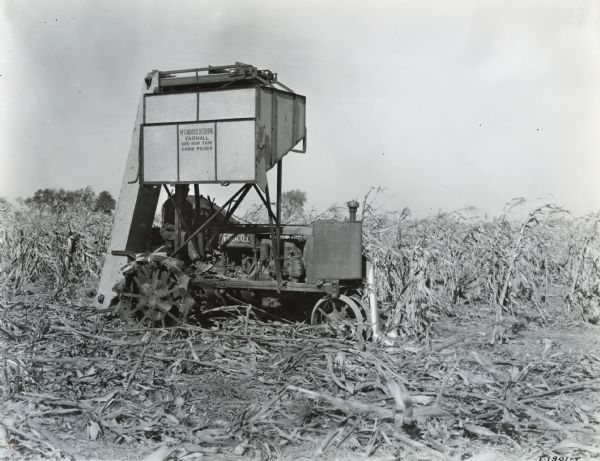 Man operating a McCormick-Deering Farmall one row tank corn picker in a field. The picker is pulled by a Farmall Regular tractor. Decals and/or stencils are on the machine. Original caption reads "farm of Mr. Froman Thomasburg, Illinois."