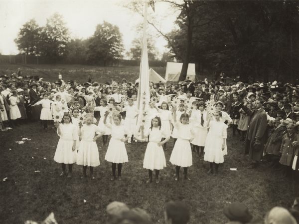 Group of girls holding fabricated flowers around a maypole at an annual school festival in Belwood Park. A large group of fellow students and teachers look on.