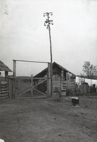 View of barnyard of crude shacks, a clothesline with clothing, a boiling cauldron, and a washtub with washboard set on a log. Ceramic jugs with holes in them are suspended from a pole for what appears to be birdhouses.