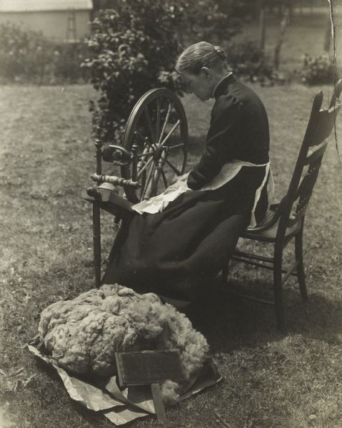 Woman sitting outdoors at a spinning wheel, turning a large pile of wool in the foreground into yarn.