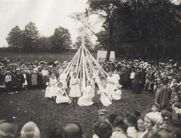 Large group of students and teachers gathered in a circle to watch young girls winding the maypole at an annual school festival in Belwood Park.