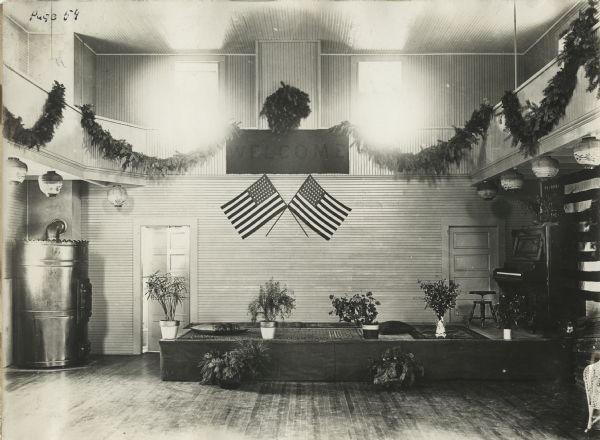 Interior of the gymnasium at Woldale Rural School, including American flags, a small stage, an upright piano and Christmas garland. In the back corner is a wood-burning stove.