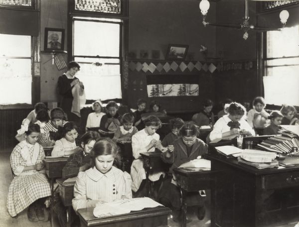 Female students working in a sewing class taught by Miss Catherine Mulvey. Original caption reads, "Miss Mulvey teaches the primary grades, but on Friday afternoons she dismisses her classes and gives a Sewing Lesson to the girls of the upper grades, while the upper grade teachers work for the boys.