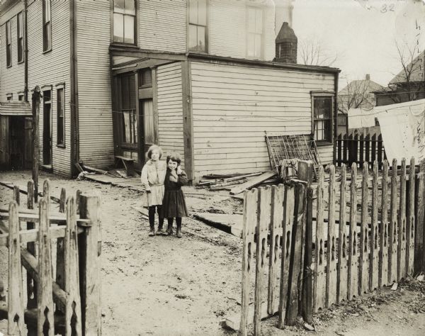 Two young girls standing with their arms around each other in the debris-strewn yard of a run-down home. The original caption identifies the location as the Ed Bauer home at the corner of Crane and Terry Streets in East Dayton.