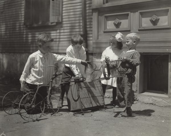 Three boys and one girl playing outside Sunnyside School with a tricycle, toy wagon, and a twisted iron chair.