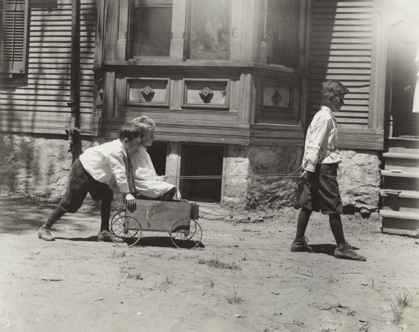 Young children playing with a toy wagon. An older boy is pulling a young girl sitting in a chair in a wagon, while another boy is pushing from behind. The group is outside Sunnyside School.