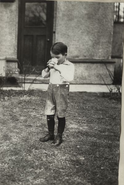 Young boy drinking milk(?) from a glass while standing outdoors. A building or house is in the background.