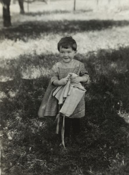 Smiling young girl standing in a yard holding fabric, perhaps an apron.