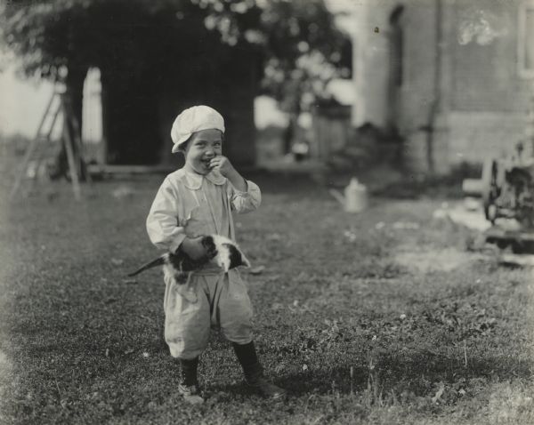 Young child standing outdoors on a lawn holding a kitten under one arm.
