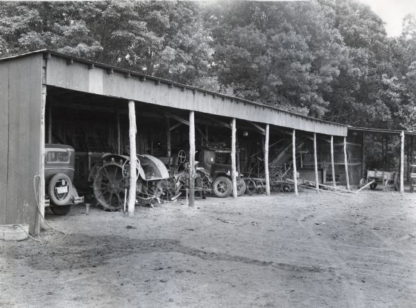 Automobiles and farm equipment are housed within a machine shed on an International Harvester demonstration farm.