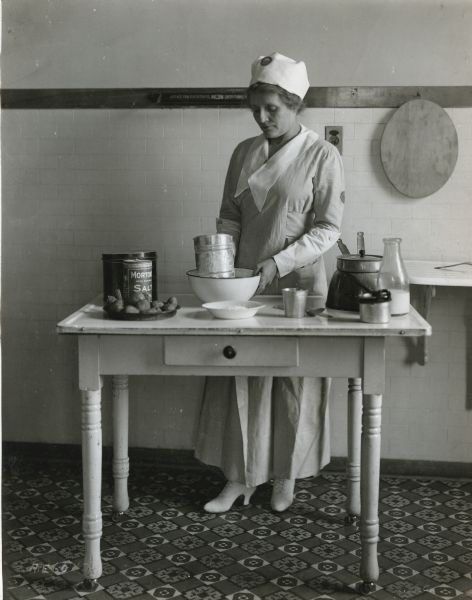 A woman wearing a uniform and cloth hat is kneading bread dough on a board on a wooden table. Her dress has buttoned-on cuffs on the sleeves. A flour sifter, bowl, and loaf pan are sitting on the table.