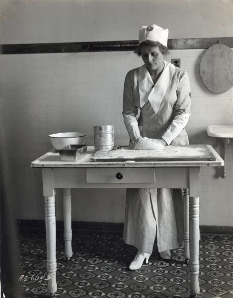 A woman wearing a uniform and cloth hat is siftting flour into a bowl in a kitchen. Her dress has buttoned-on cuffs on the sleeves. The table in front of her is lined with baking supplies, including a bottle of milk, measuring cups, salt, and potatoes.