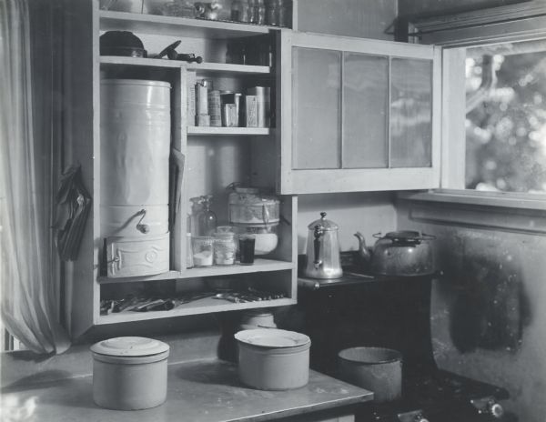 View of a corner of a kitchen with an opened window above a stove top. The kitchen is equipped with cupboards that are lined with spices, containers, and other cooking supplies above a counter.