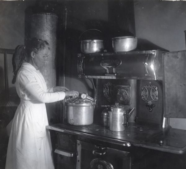 A woman wearing a full-length apron using a pressure kettle on a stove top, probably for home canning.