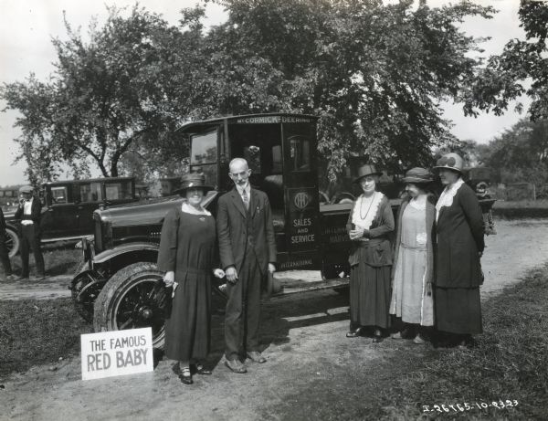 A group of women, and one man, are standing beside a McCormick-Deering Red Baby Speed Truck. The truck had a 1,500 pound capacity and was also known as the Model S. Other individuals are standing near another automobile in the background.