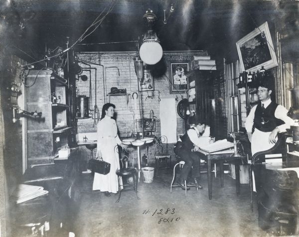A woman is holding a metal case as a man is standing and leaning against a desk in what appears to be a medical office or infirmary. The office is most likely at International Harvester's McCormick Works. Another man is sitting at a drafting table to the right; a cot is on the left, and framed artwork, including a McCormick poster, decorates the walls.