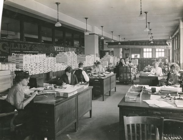 Men and women are working at desks in an International Harvester office as other employees unload packages from what appears to be a mail cart in the background. Advertisements for Lily cream separators and Weber wagons are hanging on the wall to the left. Stacks of pamphlets, possibly advertising brochures, are on the left. The offices may be part of the Harvester Press operations.