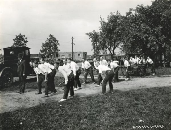 A group of men, possibly fire fighters, appear poised to take part in a hook and ladder competition. A Red Baby truck and other vehicles are parked nearby. One man in the foreground appears to be holding a fire hose.
