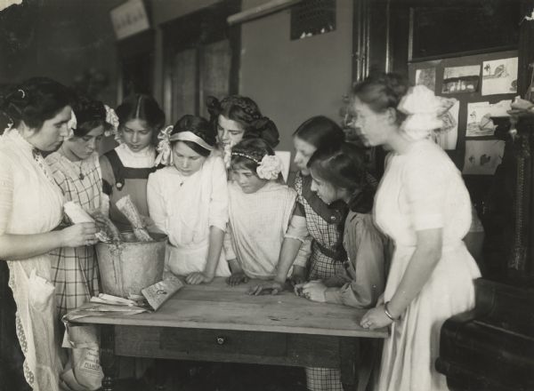 Class of girls and teacher running a seed germination test ("rag-doll" test) in the classroom. The class is standing around a table, and are using a pail to test the corn which is wrapped in newspaper bundles.