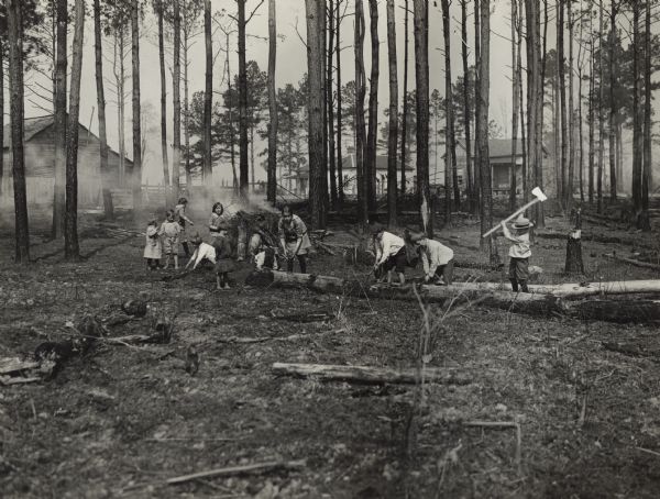 Several children clearing land for a new school playground. Three boys are using axes to chop a log, and other children are gathered near the fire. Three buildings are in the background beyond a stand of trees. Original caption reads: "A few second and third grade school boys and girls at work clearing a play ground at Lisman, Ala. These children are doing this of their own free will and without an older person to direct their work. From the looks of the burning pile of brush they seem to have made good headway."