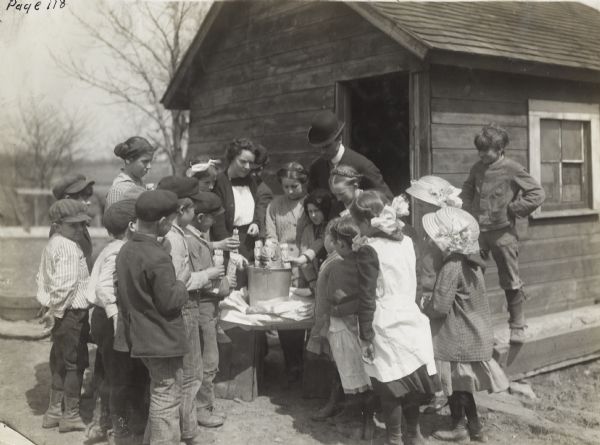 Class of children gathered outdoors around a table for a corn germination lesson. Original caption reads: "after the rag dolls are prepared they are immersed in water for a few minutes and then packed in buckets till germination takes place. School children working under the direction of Prof. Tobin of Cook County."