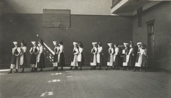 High school girls in a folk dancing class, probably in the school gymnasium. The girls are in a line grouped in pairs, and are wearing matching dresses and hats. A basketball hoop is in the background, and there is a piano is the back right corner.