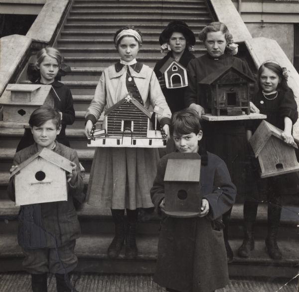 Group of school children standing on the steps of a school house(?) holding their bird house projects.